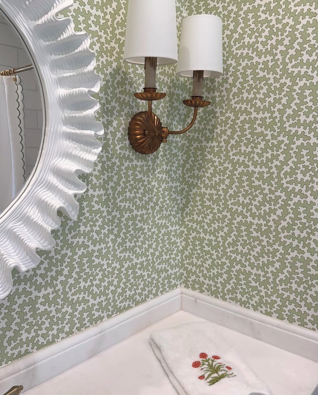 Dover Apple clay coated wallpaper with a subtle green abstract pattern, a white ruffled mirror, and brass sconce.