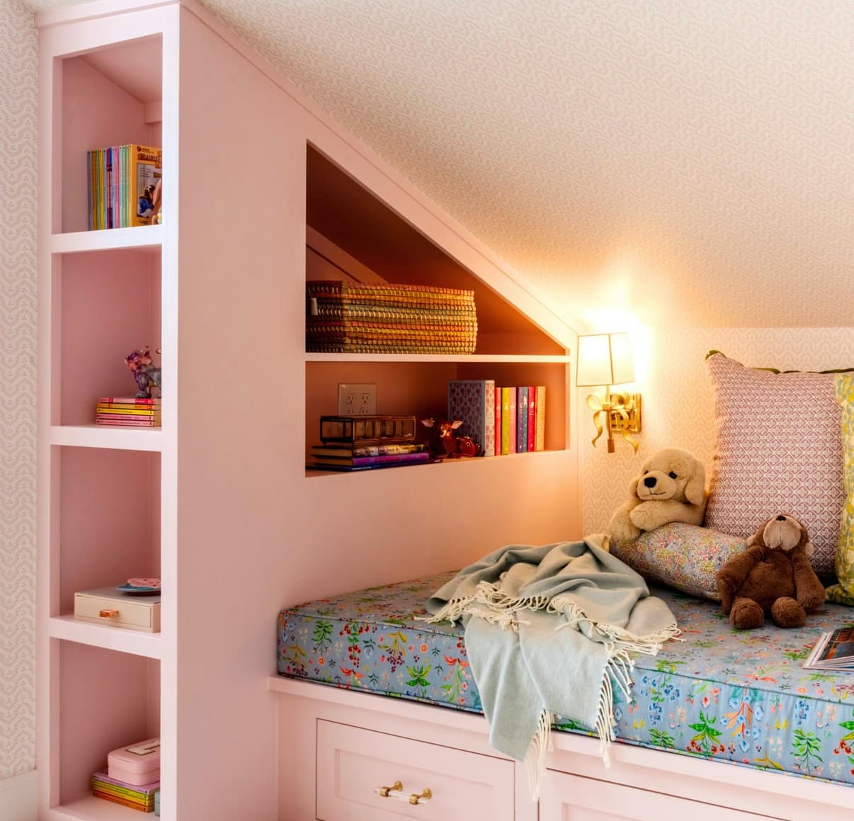 Pink built-in shelves and window seat with blue floral fabric cushion, stuffed animals, and books.