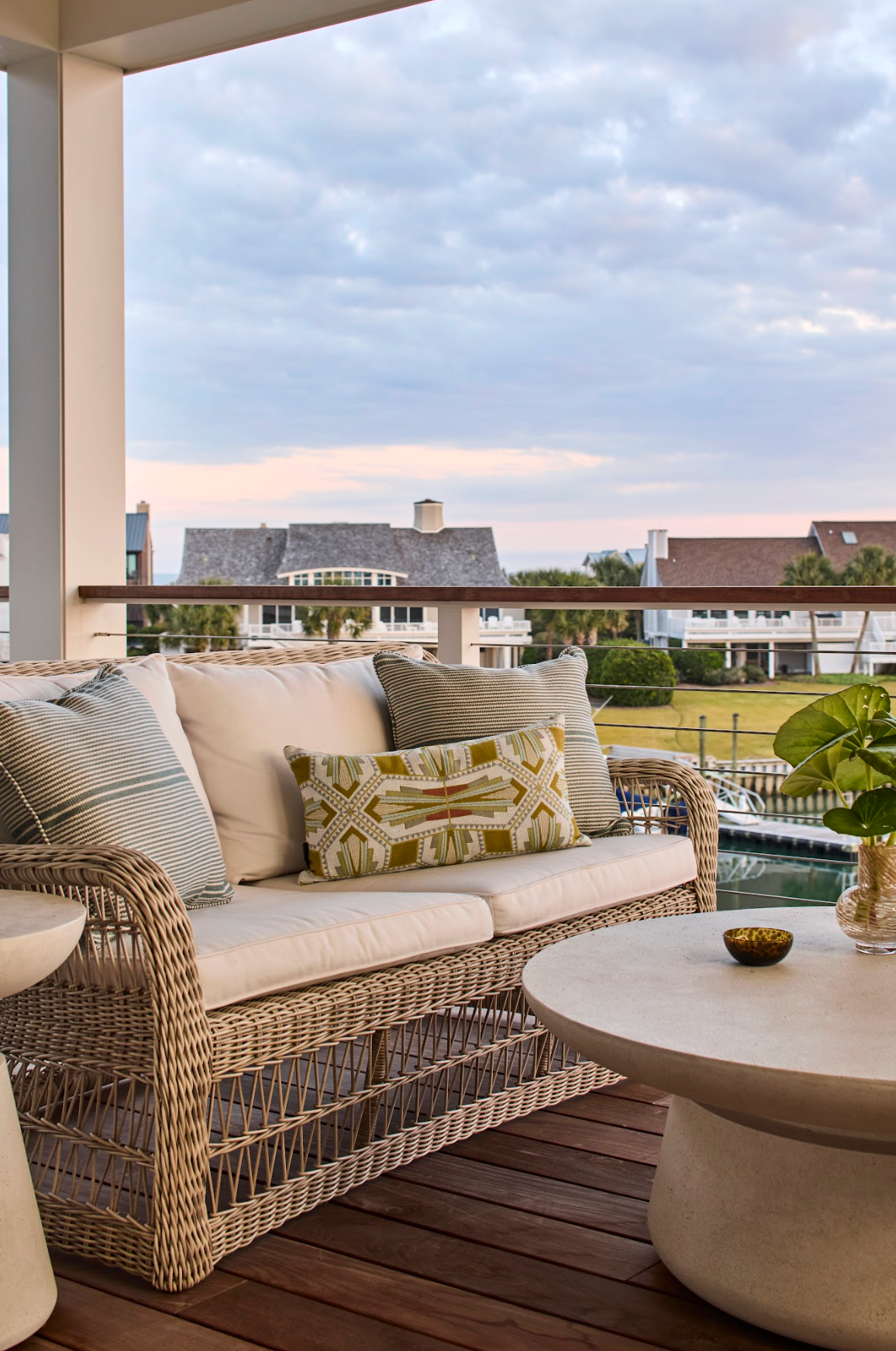 Woven wicker outdoor sofa with cream cushions and decorative pillows on a deck overlooking houses and water.