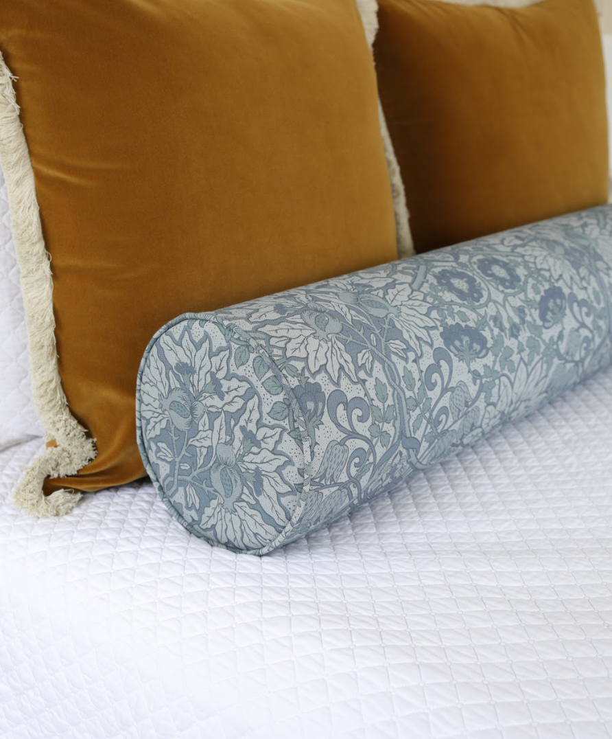 Cobalt Flora Fabric bolster pillow with a botanical pattern, next to two mustard velvet pillows on a white quilted bed.