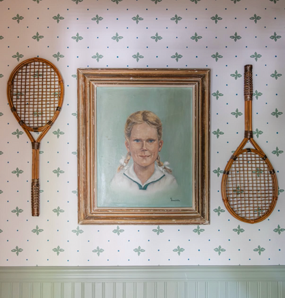 Folly Fern wallpaper with green botanical pattern, framing a vintage portrait and two rattan tennis rackets.