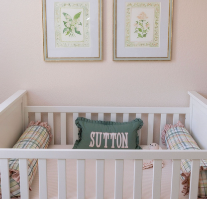 White crib with pink striped sheet, green "SUTTON" pillow, and two plaid bolster pillows.