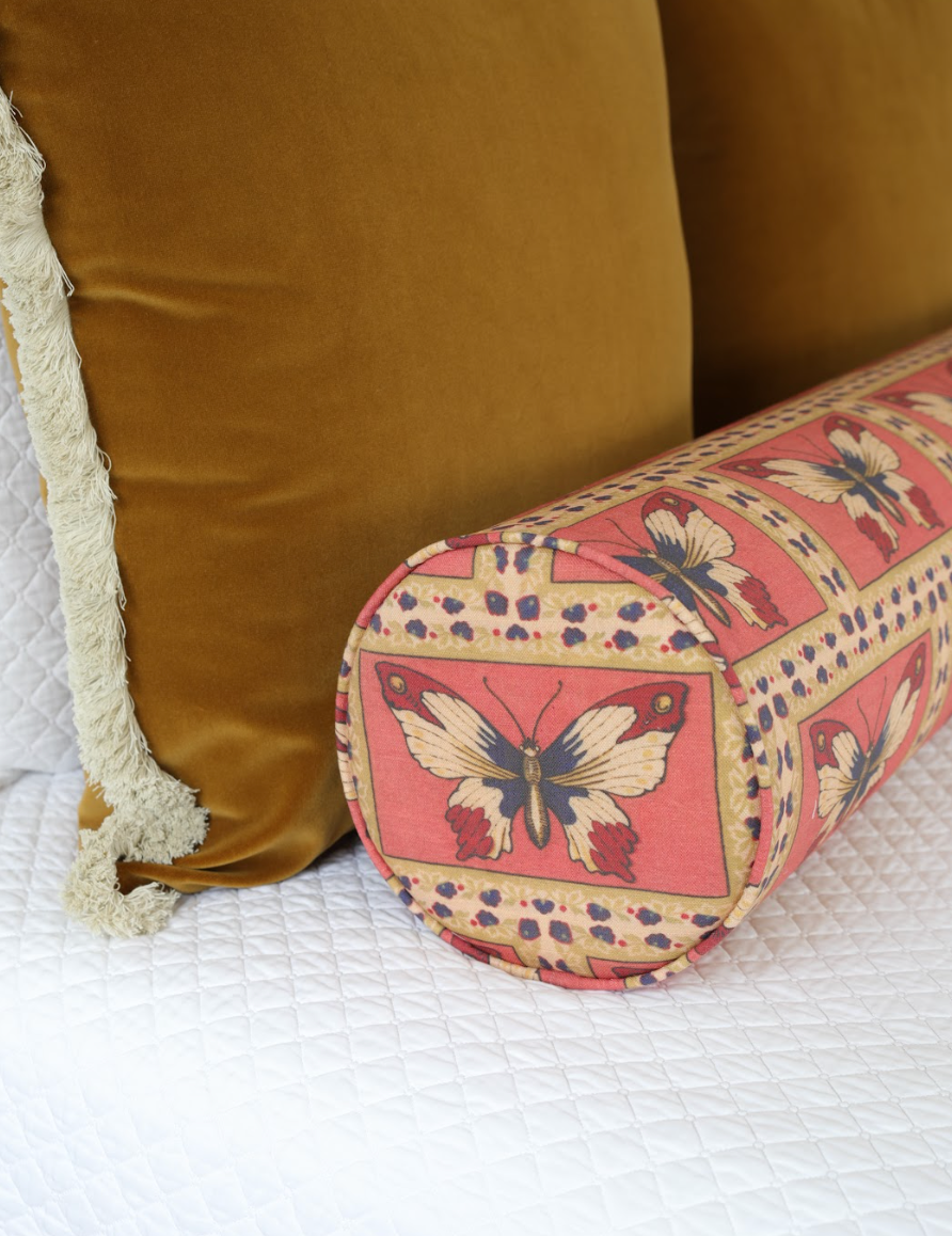 Monarch Fabric - Rouge, a linen-cotton blend with butterfly pattern, on a bolster pillow next to a gold velvet pillow.