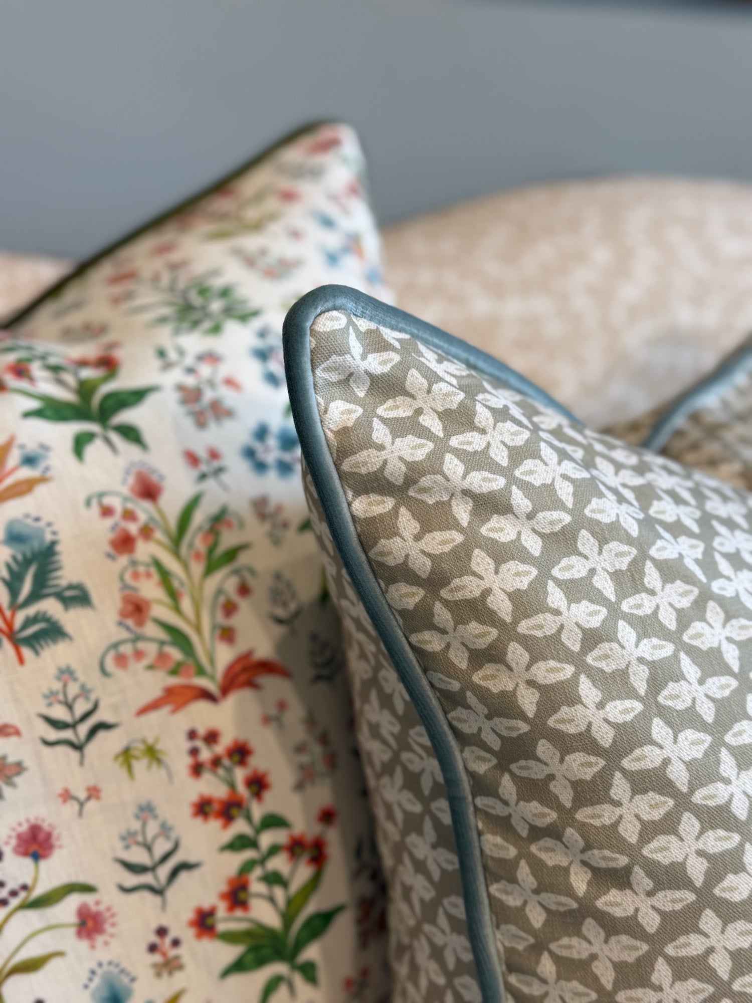 Close-up of two linen-cotton blend throw pillows. Front pillow is sage green with white floral pattern and blue piping.