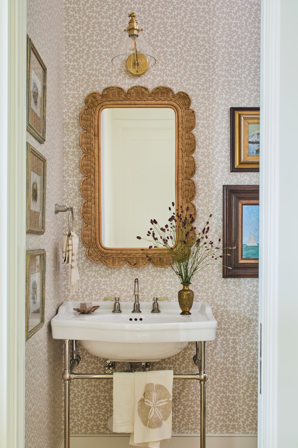 Dover Wallpaper in Sand with a subtle floral repeat, framing a wicker mirror and pedestal sink in a bathroom.
